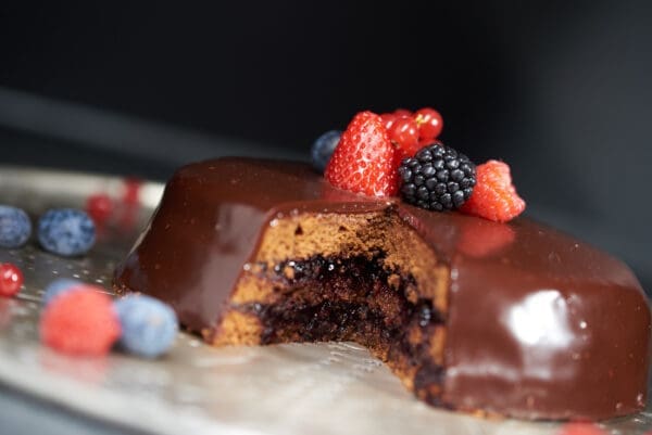 A chocolate-glazed cake, featuring a slice removed to show a dark filling enriched with Luxardo Raspberry Jams and Spreads (4x5.4Li), is topped with fresh strawberries, blackberries, and blueberries. It rests on a silver platter decorated with scattered berries.