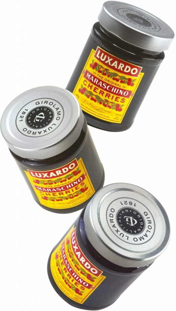 Three Luxardo Maraschino Cherries (400g jars) with red and yellow labels are arranged diagonally on a white background; two jars stand upright while one lies on its side.