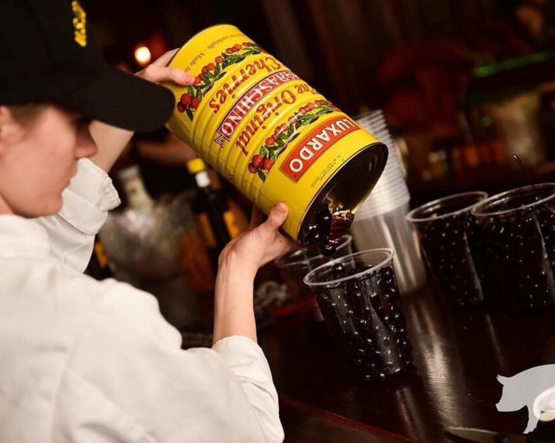 A person in a white shirt pours black olives from a large yellow can into clear plastic cups on a dark countertop.
