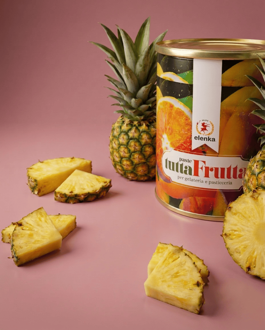 A can of Elenka TuttaFrutta sits on a white surface with whole pineapples and several pineapple slices, against a pink and light green background.