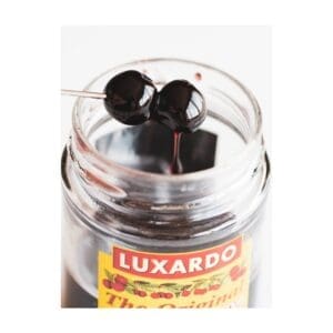 A jar of Luxardo cherries with two dark cherries on a cocktail pick above the open jar.