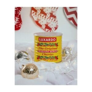 A yellow can of Luxardo maraschino cherries sits among string lights and gold ornaments, with red and white Christmas stockings hanging in the blurred background.