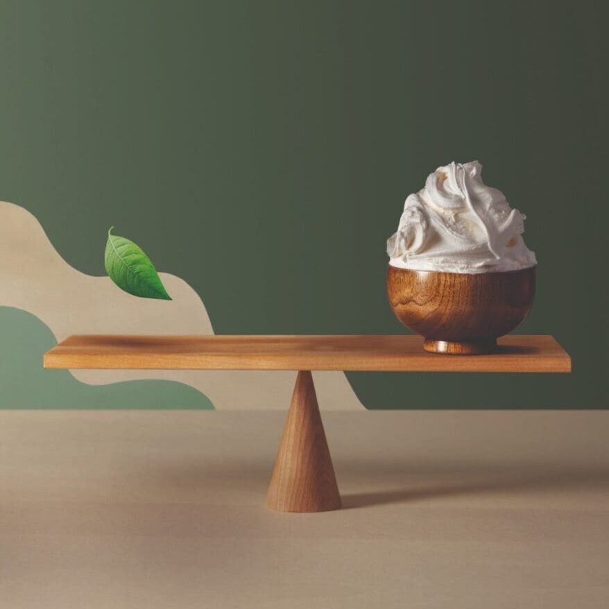 A wooden pedestal balancing a small wooden bowl filled with whipped cream. The background is a two-tone wall, green and cream, with a decorative leaf element on the green side. The pedestal has a modern, minimalist design, reminiscent of Italian products known for their elegance and craftsmanship.