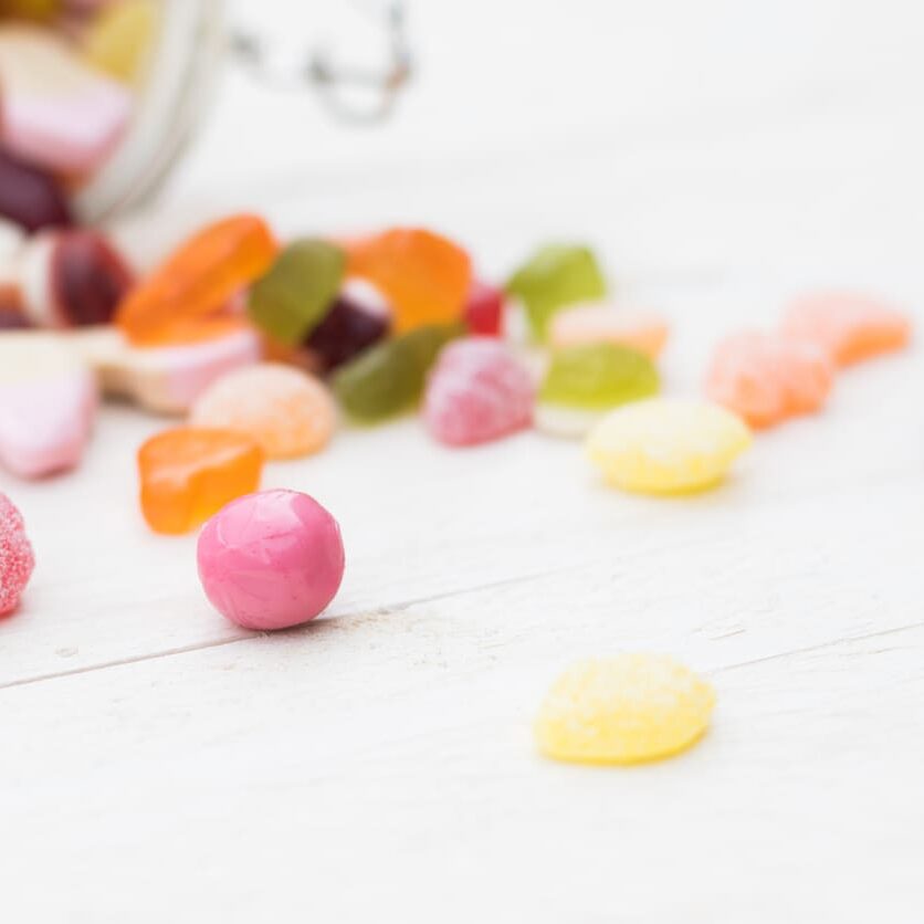 Open glass jar full of different colourful jelly candies on white wooden background