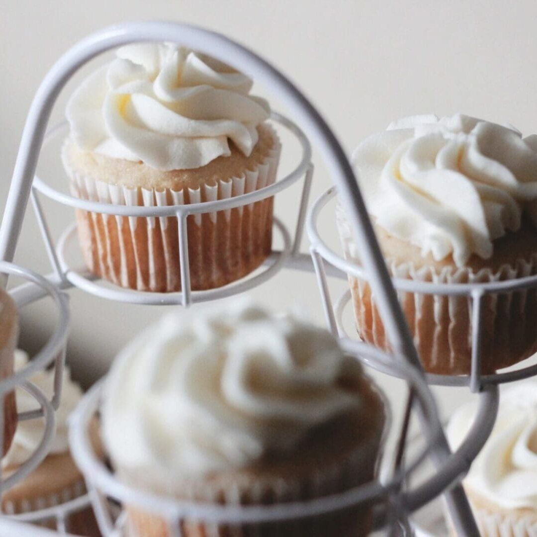 A close-up shot of several vanilla cupcakes with swirled white frosting displayed on a white metal cupcake stand. The background is soft and blurred, bringing the focus to the cupcakes’ intricate frosting details.